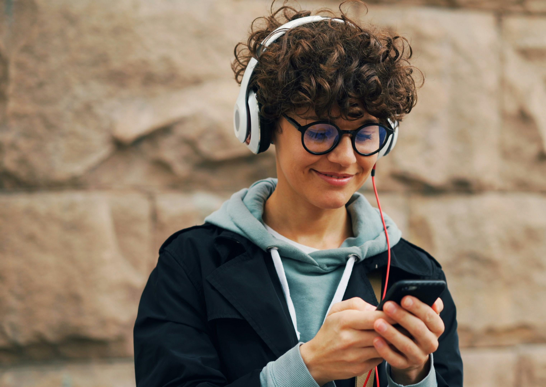 Young person smiling looking at phone while outside wearing headphones